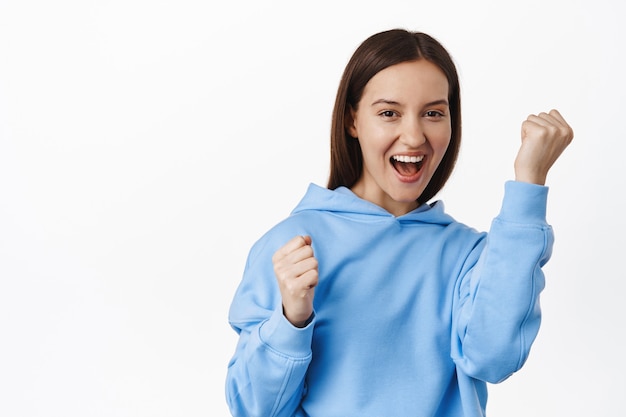 Self-assured young woman feel encouragement boost, raise fist up and smiling confident, rooting and chanting, winning and triumphing, standing against white wall.