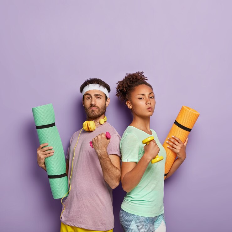 Free Photo self confident serious diverse woman and man stand back to each other, raise arms with dumbbells, hold karemats, show their strength, being in good physical shape, isolated on purple  wall