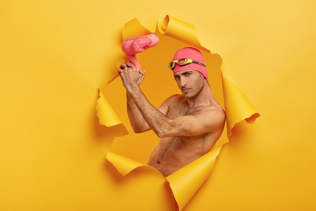 Free photo self confident serious guy with bristle holds pink inflated flamingo, wears swimcap with goggles on forehead