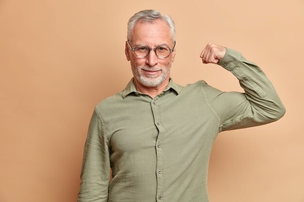 Self confident serious man raises arm shows muscles being confident in his strength wears formal shirt poses against brown wall