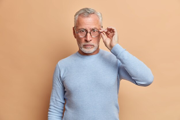 회색 수염을 가진 자기 확신 진지한 남자는 안경에 손을 유지합니다. 캐주얼 점퍼를 입은 정면에 직접 보이는 정보는 신중하게 베이지 색 벽에 포즈를 취합니다.