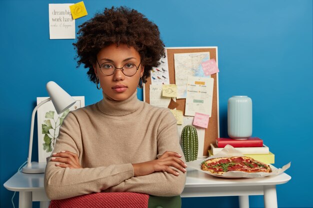 Self confident serious woman with Afro hair, keeps hands crossed, leans at chair, works at scientific project at home, has break for snack and rest, poses against desktop with books, notes, lamp