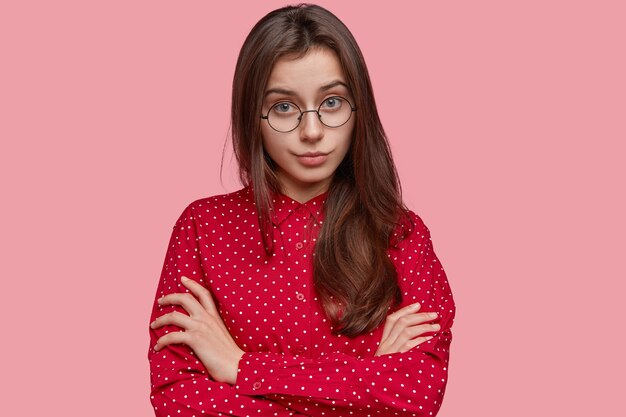 Self confident serious young lady keeps arms folded, dressed in polka dot outfit, looks attentively directly , wears round glasses
