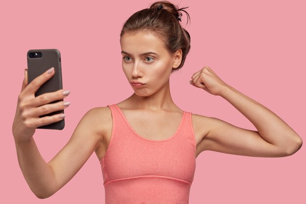 Self confident woman with dark hair, shows muscles, raises hand, makes selfie portrait