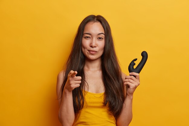 Self confident young woman points index finger, holds vibrator to stimulate clitoris with scintillating vibrations, has personal dildo, isolated on yellow wall. Sex toy for women.