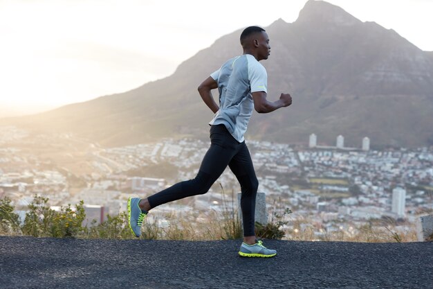 自己決定の暗い肌のスポーティな男性ランナーは、スポーツ服を着て、山道を遠距離恋愛し、新鮮な空気を楽しみ、エネルギッシュでやる気を感じます。人、ライフスタイル、スポーツのコンセプト