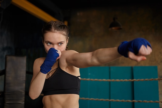 Self determined young woman professional boxer with strong muscular arms and abdominal wearing black sports top and blue boxing bandages mastering punching technique in gym