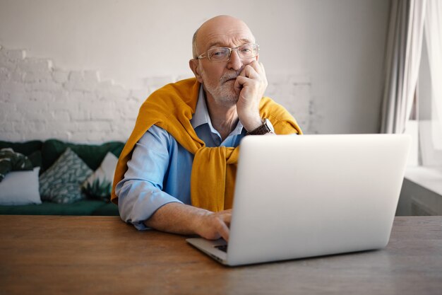 自営業の無精ひげを生やした老人がポータブルコンピュータでキーボード操作を行い、遠隔作業にワイヤレスインターネット接続を使用しています。オンラインコミュニケーションを楽しんでいる眼鏡の現代のシニア成熟した男性
