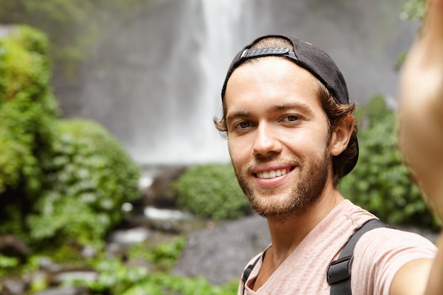 Self-portrait of happy hiker in baseball cap taking selfie while standing against waterfall in green exotic woods. Young tourist trekking in rainforest during his holidays