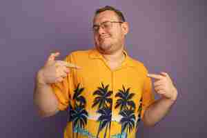 Free photo self-satisfied man in glasses wearing orange shirt pointing at himself happy and cheerful standing over purple wall