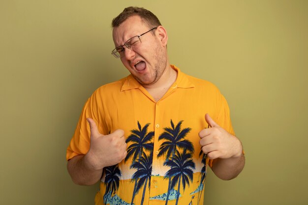 Free photo self-satisfied man in glasses wearing orange shirt  pointing at himself smiling and winking standing over green wall