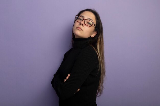 Self-satisfied young beautiful girl in a black turtleneck and glasses lookign at camera with crossed arms on chest 