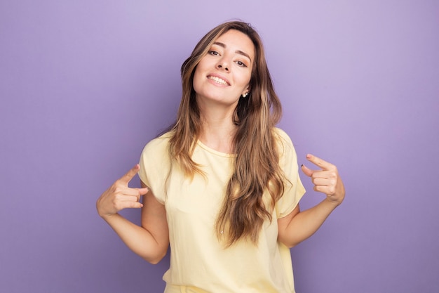 Самодовольная молодая красивая женщина в бежевой футболке выглядит уверенно улыбаясь, указывая на себя, стоящую над фиолетовым