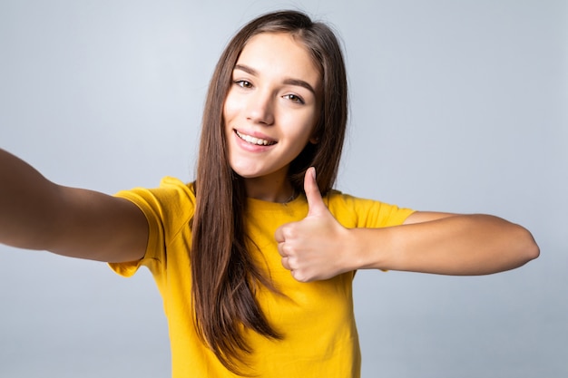 Selfie of happy woman showing thumb up and winking isolated on white wall
