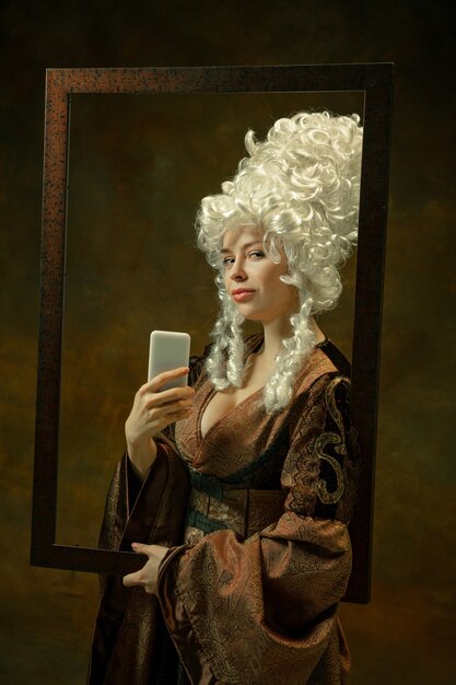 Selfie in mirror. Portrait of medieval woman in vintage clothing with wooden frame on dark background. Female model as a duchess, royal person. Concept of comparison of eras, modern, fashion, beauty.