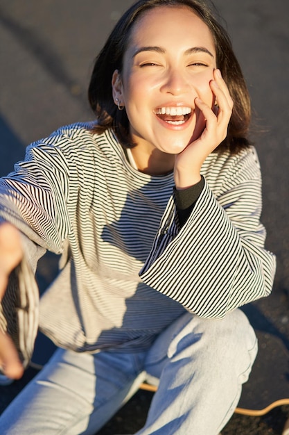 無料写真 屋外のスケート ボードに座ってスマート フォンで写真を撮る笑顔の美しいアジアの女の子の selfie