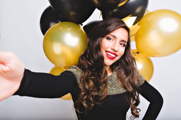 Selfie portrait funny amazing girl in elegant fashion dress between gold and black balloons on white space