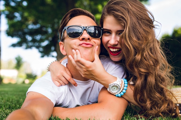 Selfie-portrait of funny couple  lying on grass in summer park. Girl with long curly hair, red lips and young guy in sunglasses aping to camera.