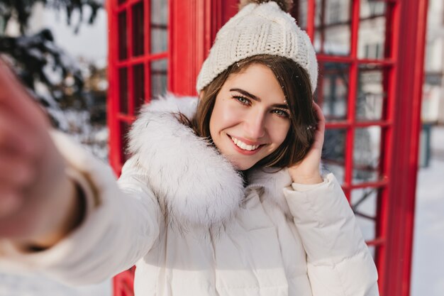 Selfie portrait joyful pretty woman in white woollen hat enjoying sunny winter morning on red telephone box