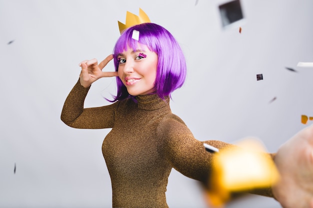 Selfie portrait pretty woman with violet haircut having fun in gold tinsel. She has gold crown, smiling
