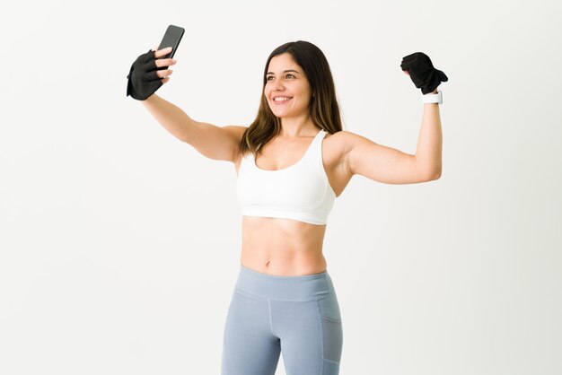 Selfie to show off my progress at the gym. Beautiful happy woman smiling while taking a picture with her smartphone and showing her strong biceps