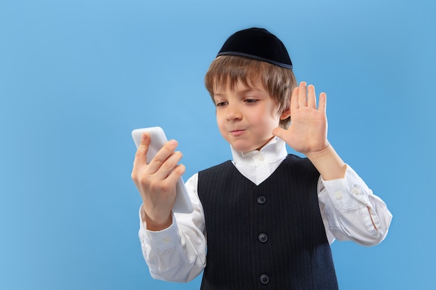 Selfie, vlog. Portrait of a young orthodox jewish boy isolated on blue wall. Purim, business, festival, holiday, childhood, celebration Pesach or Passover, judaism, religion concept.