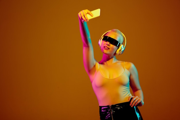 Selfie. Young caucasian woman on brown  wall in neon light. Beautiful female model with fashionable, trendy eyewear. Human emotions, facial expression, sales, ad concept. Freak's culture.