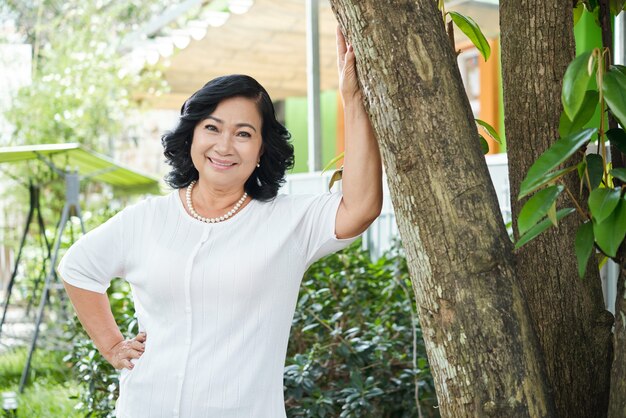 Senior Asian woman posing in garden and leaning on tree