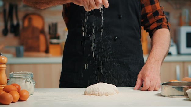 Senior bakery man sifting wheat flour on dough for kneading. Retired elderly chef with bonete and uniform sprinkling, sieving spreading rew ingredients with hand baking homemade pizza and bread.