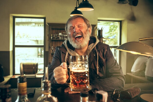 Senior bearded man drinking alcohol in pub and watching a sport program on TV. Enjoying my favorite teem and beer. Man with mug of beer sitting at table. Football or sport fan. Human emotions concept