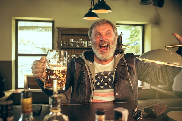 Senior bearded man drinking alcohol in pub and watching a sport program on TV. Enjoying my favorite teem and beer. Man with mug of beer sitting at table. Football or sport fan. Human emotions concept