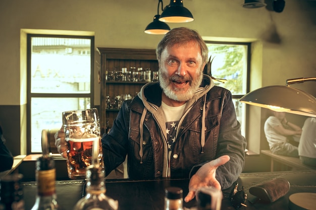 Senior bearded man drinking alcohol in pub and watching a sport program on TV. Enjoying my favorite teem and beer. Man with mug of beer sitting at table. Football or sport fan. Human emotions concept