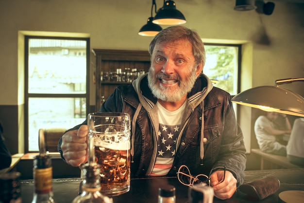 Foto gratuita uomo barbuto anziano che beve alcolici in un pub e guarda un programma sportivo in tv. gustando il mio brulicare e la mia birra preferiti. uomo con boccale di birra seduto a tavola. appassionato di calcio o di sport. concetto di emozioni umane