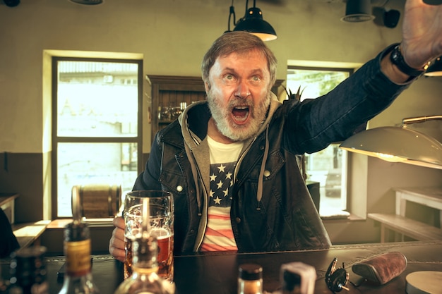 Uomo barbuto anziano che beve alcolici in un pub e guarda un programma sportivo in tv. gustando il mio brulicare e la mia birra preferiti. uomo con boccale di birra seduto a tavola.