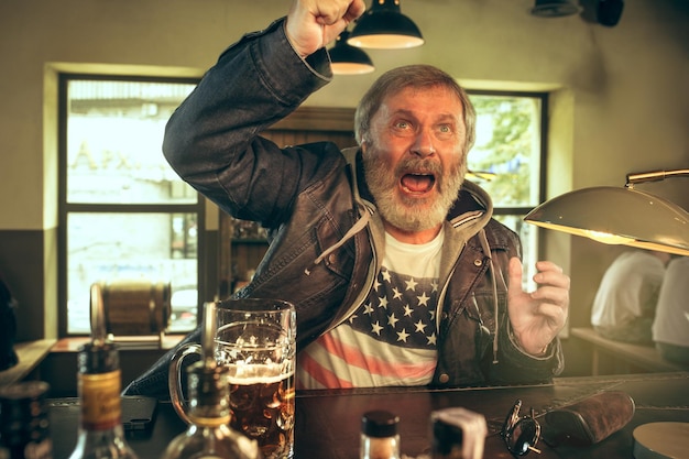 Senior bearded man drinking alcohol in pub and watching a sport program on TV. Enjoying my favorite teem and beer. Man with mug of beer sitting at table. 