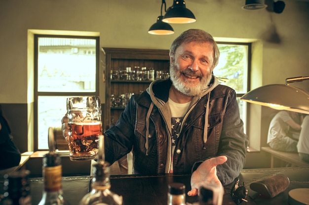 Uomo barbuto anziano che beve alcolici in un pub e guarda un programma sportivo in tv. gustando il mio brulicare e la mia birra preferiti. uomo con boccale di birra seduto a tavola.