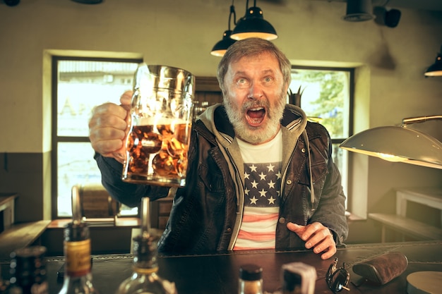 Uomo barbuto anziano che beve alcolici in un pub e guarda un programma sportivo in tv.