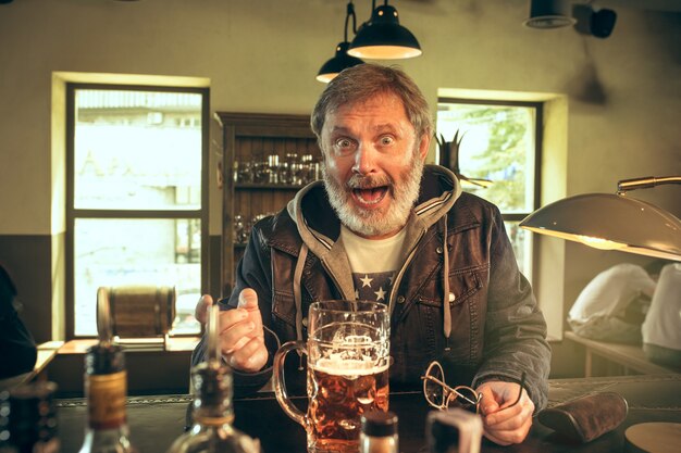 Foto gratuita uomo barbuto anziano che beve alcolici in un pub e guarda un programma sportivo in tv.