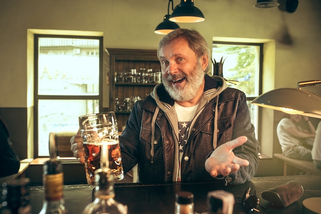 Senior bearded man drinking alcohol in pub and watching a sport program on TV.