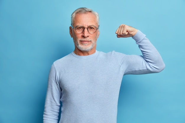 Старший бородатый мужчина показывает мышцы после занятий бодибилдингом, носит прозрачные очки и простой джемпер позирует на синей стене студии