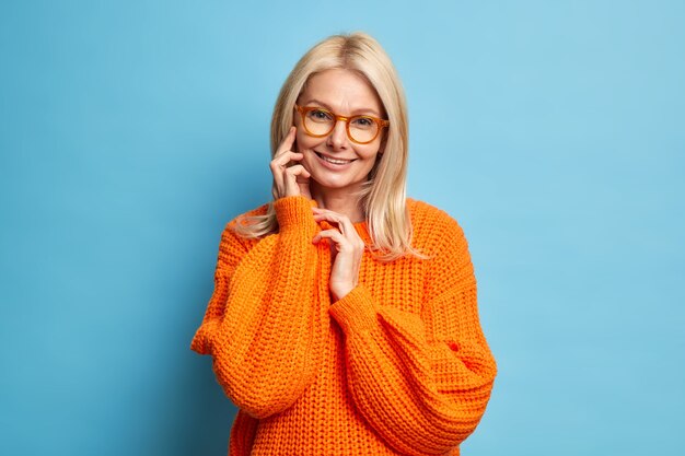 Старшая красивая женщина со светлыми волосами, здоровая кожа трогает лицо, нежно улыбается, нежно носит оранжевый вязаный свитер модели