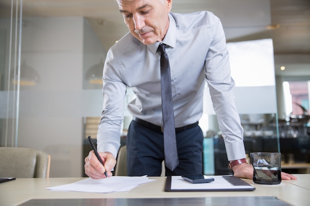 Senior Businessman Bending over Desk and Writing