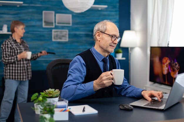 Senior businessman holding cup of coffee working on laptop. Elderly man entrepreneur in home workplace using portable computer sitting at desk while wife is holding tv remote.