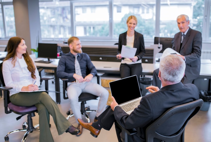 Free Photo senior businessman sitting on chair with laptop sitting in front of his team in the office