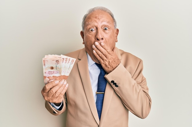 Senior caucasian man holding 10 colombian pesos banknotes covering mouth with hand shocked and afraid for mistake surprised expression
