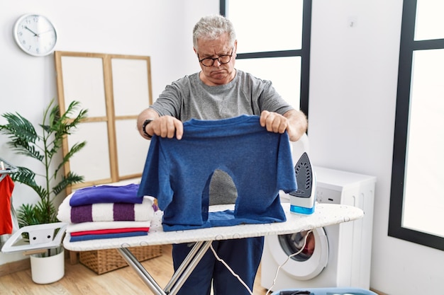 Free photo senior caucasian man ironing holding burned iron shirt at laundry room relaxed with serious expression on face. simple and natural looking at the camera.