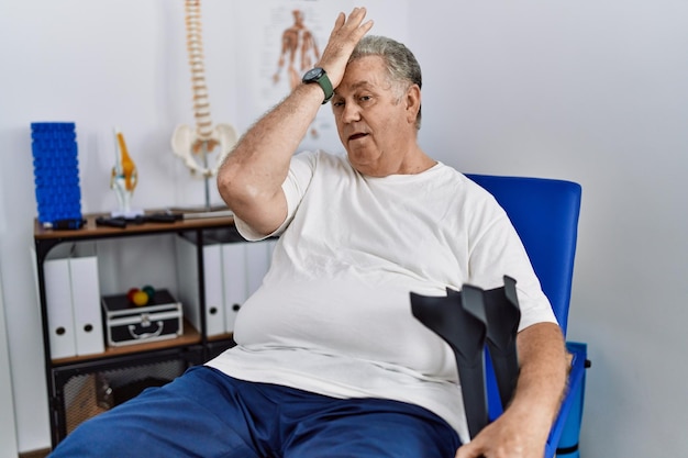 Free photo senior caucasian man at physiotherapy clinic holding crutches surprised with hand on head for mistake, remember error. forgot, bad memory concept.