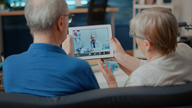 디지털 태블릿으로 의사와 온라인 회의에 참석하는 노인 부부는 집에서 원격 상담을 하고 있습니다. 인터넷 원격 의료를 위해 현대적인 기기에서 화상 회의를 사용하는 노인들.