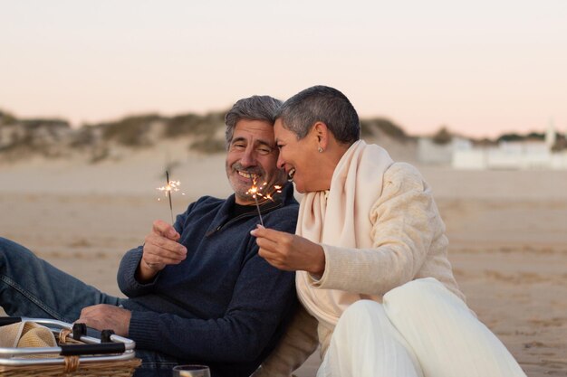 Senior couple enjoying picnic at seashore, holding sparklers and laughing. Lady with short hair and happy grey-haired man having fun together at sunset on the beach. Love, relationship concept