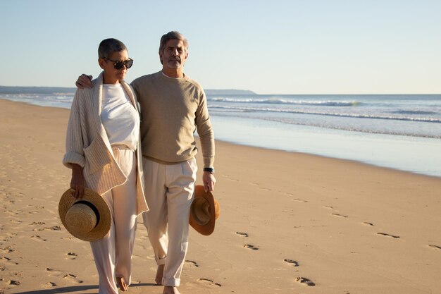 Пожилая пара обнимается, прогуливаясь в одиночестве по берегу моря. Красивый бородатый мужчина и короткошерстная дама в солнцезащитных очках с соломенными шляпами, наслаждаясь отдыхом на пляже. Досуг, концепция отношений